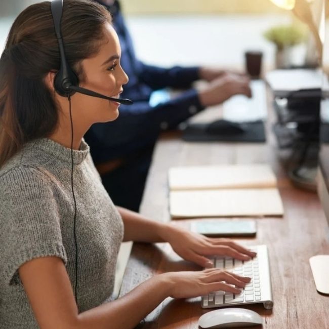 Eine nett lächelnde Frau tippt auf einer Tastatur und hat ein Headset auf. Sie trägt ein Strick-T-Shirt. Neben ihr erkennt man im Hintergrund eine weitere Person, die am PC arbeitet.