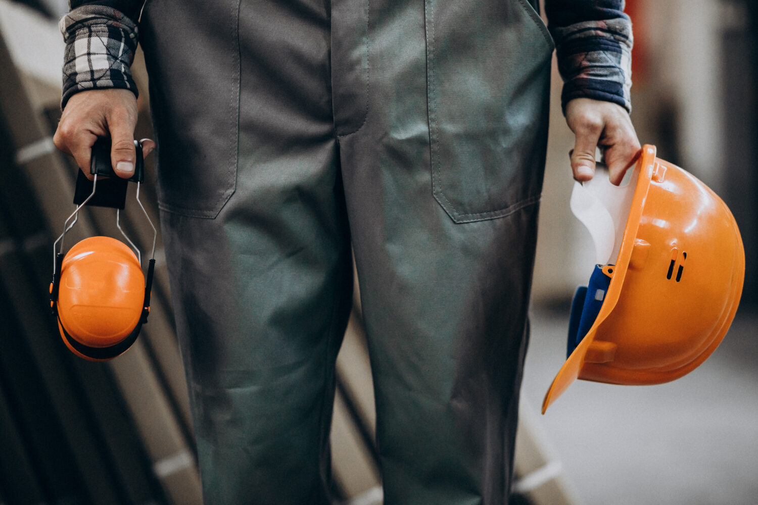 Ein Mitarbeiter hält einen orangenen Schutzhelm und Gehörschutz in den Händen. Wir sehen nur seine hängengelassenen Arme und die Beine.