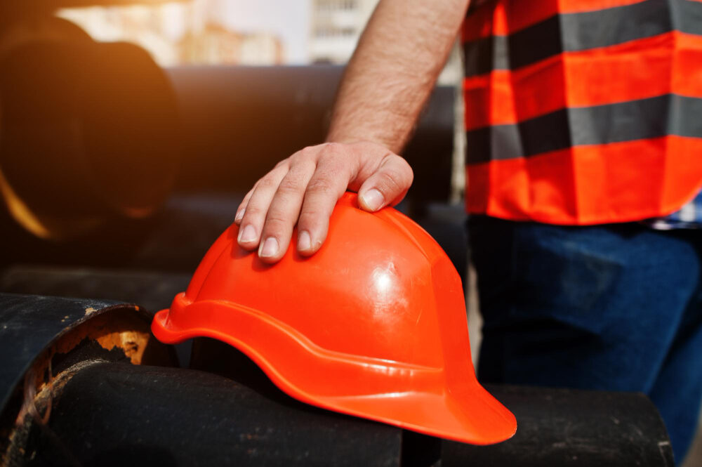 Eine Fachkraft für Arbeitssicherheit in orangener Warnweste legt eine Hand auf einen Schutzhelm, der vor ihr liegt.