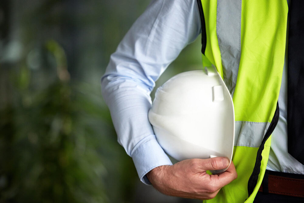 Eine Fachkraft für Arbeitssicherheit hält einen Helm unter den Arm geklemmt und trägt eine gelbe Warnweste.