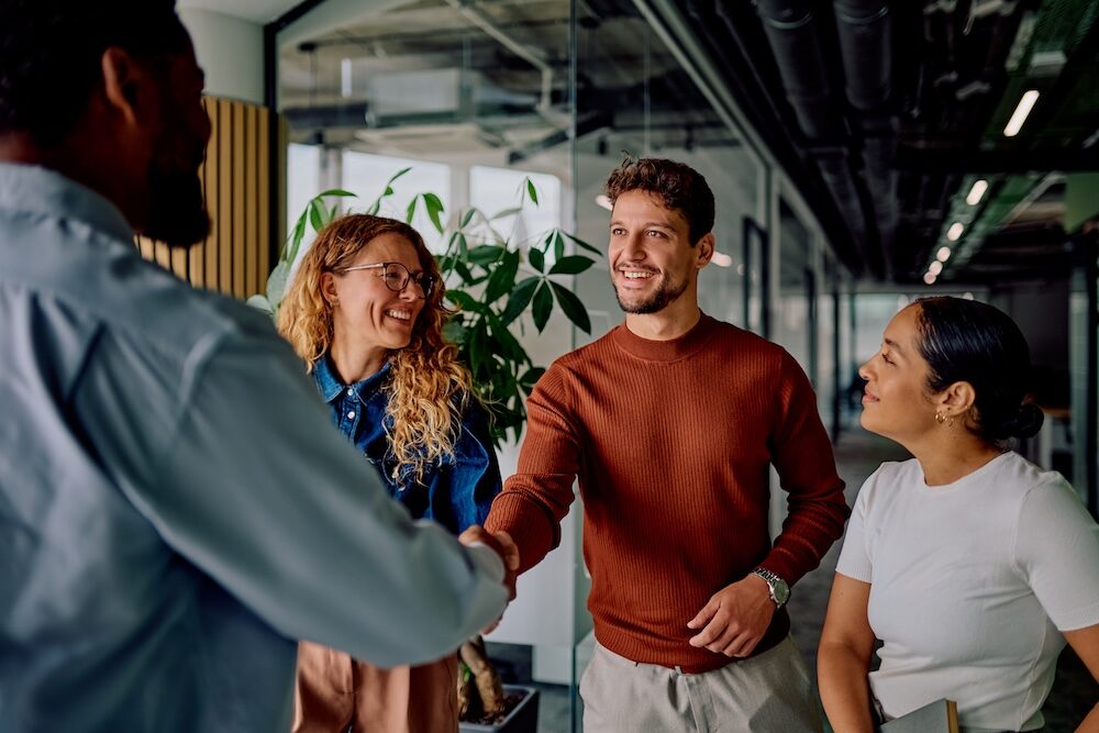 Vier glückliche Mitarbeitende lachen sich an und geben sich die Hand in einem Büro.