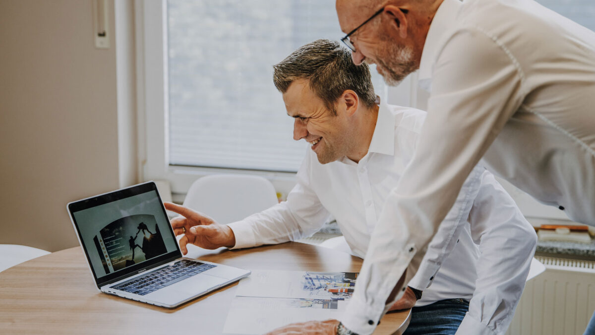 Zwei Männer reden an einem Laptop im Büro über die digitale Gefährdungsbeurteilung.