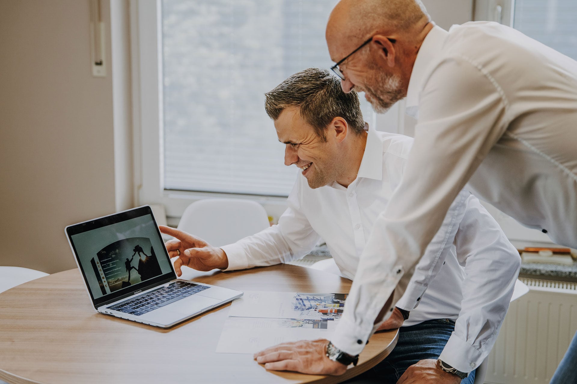 Zwei Männer reden an einem Laptop im Büro über die digitale Gefährdungsbeurteilung.